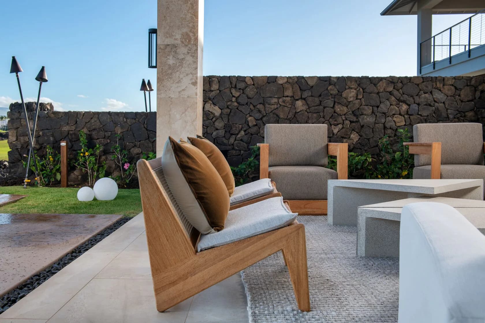Contemporary outdoor patio with wooden lounge chairs, gray cushions, stone walls, and modern furnishings designed by Rddhima Golyan.