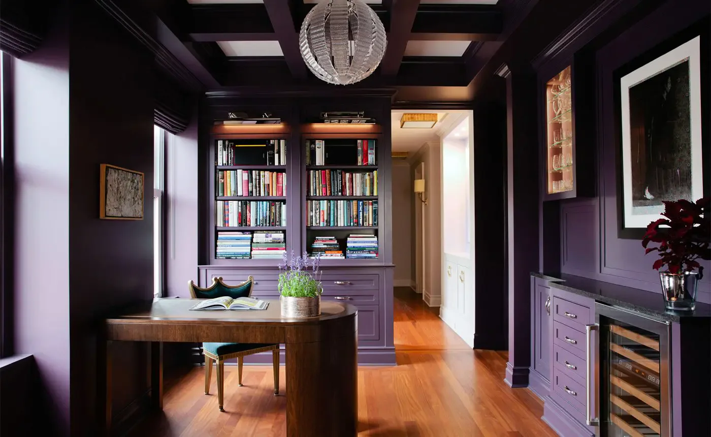 Dark purple home office with built-in bookshelves, wooden desk, hardwood floors, and layered lighting design by Rebecca Ascher.
