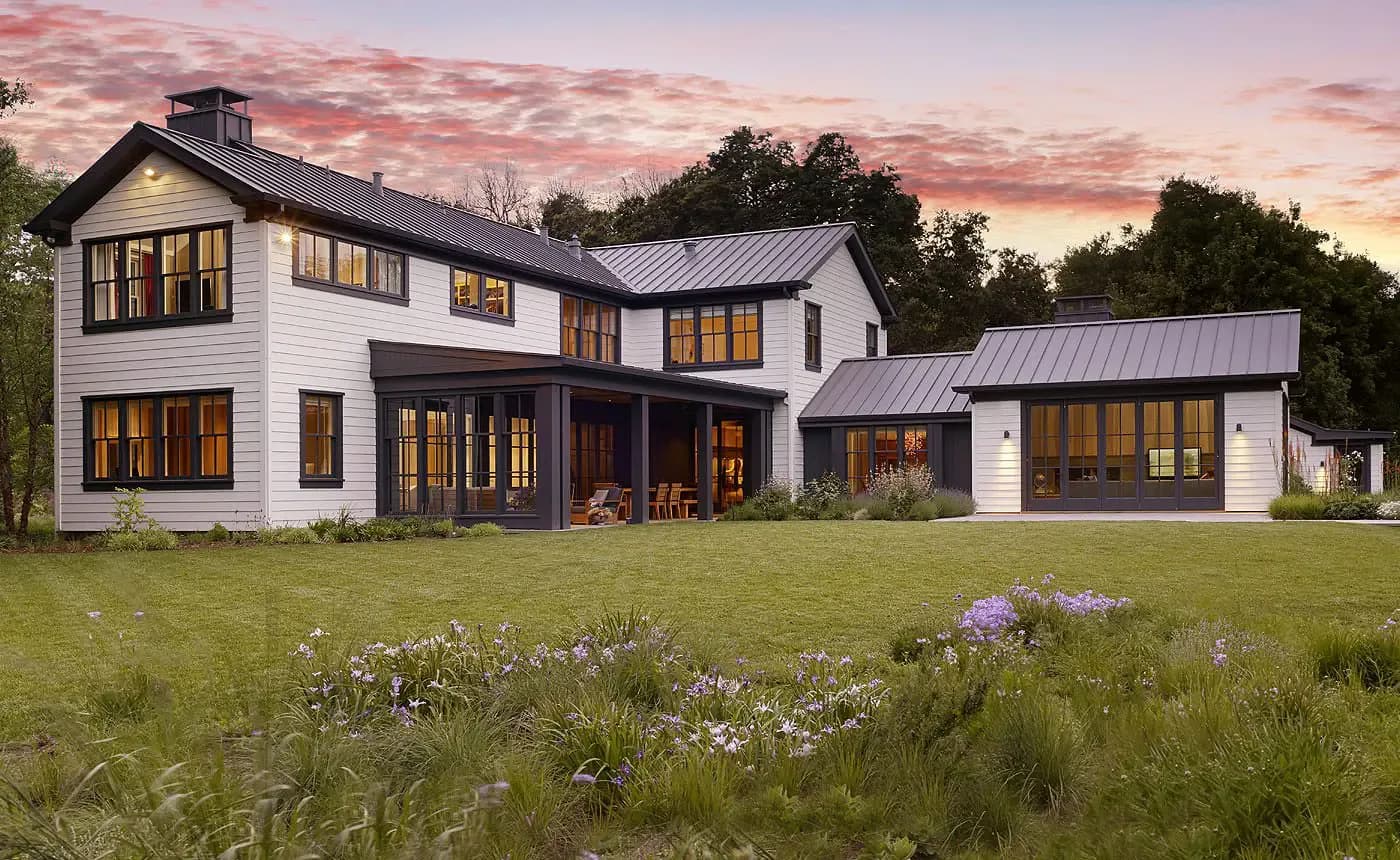 Modern farmhouse exterior with white clapboard siding, dark metal roof, and expansive windows at dusk by Rebecca Ascher.
