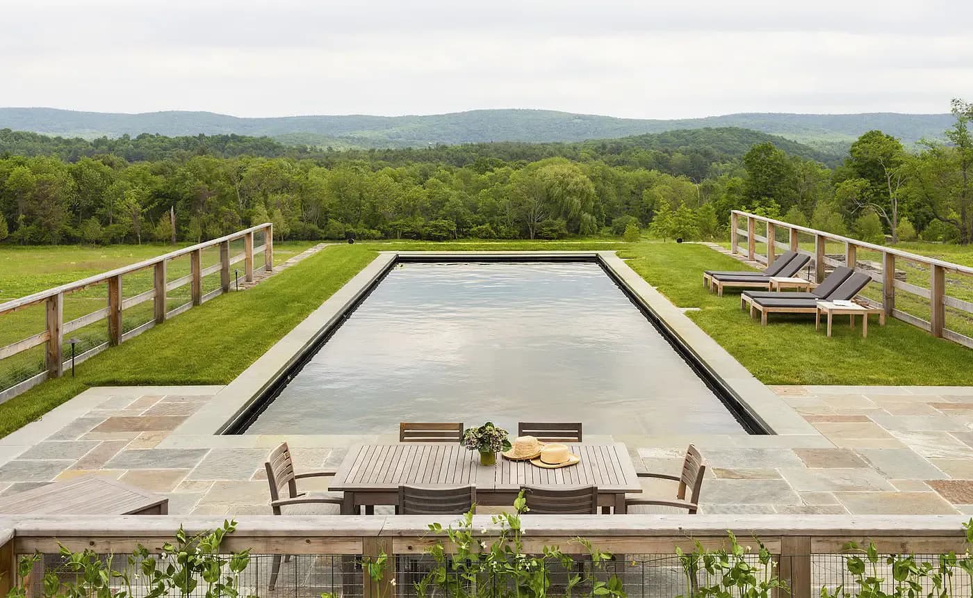 Modern rectangular swimming pool with concrete surround, wooden railings, lounge seating, and mountain landscape views, designed by Rebecca Ascher.