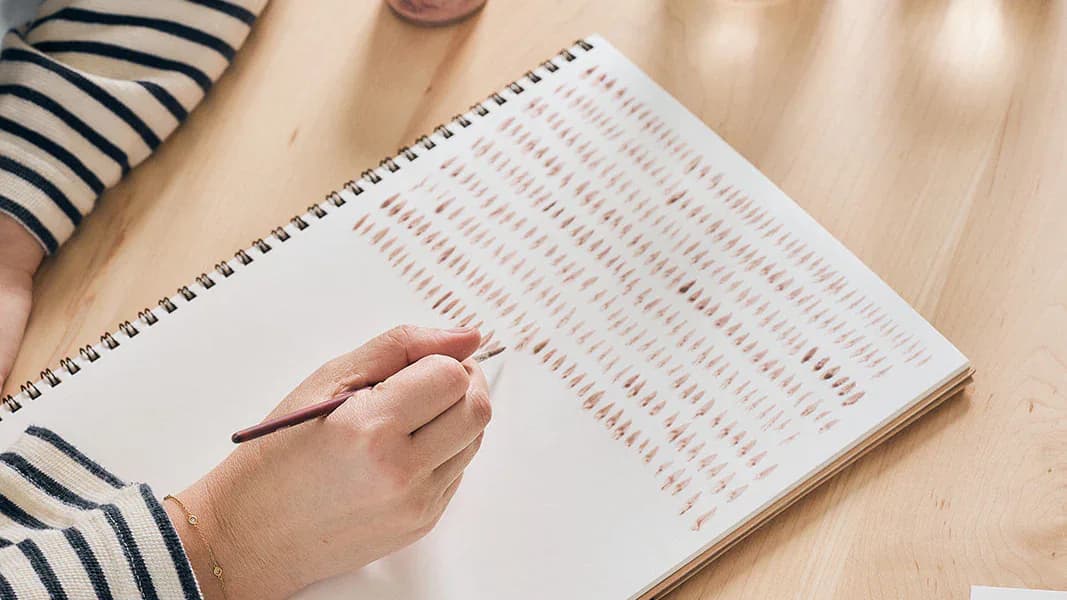 Hand holding a pen over a spiral-bound notebook with red dotted pattern on wood surface by Rebecca Atwood.