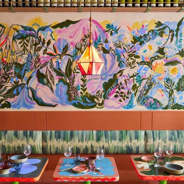 Vibrant restaurant interior featuring a large-scale floral mural in pink, blue, and yellow tones above a terracotta wall, with colorful place settings and modern pendant lighting by Rebecca Johnson.