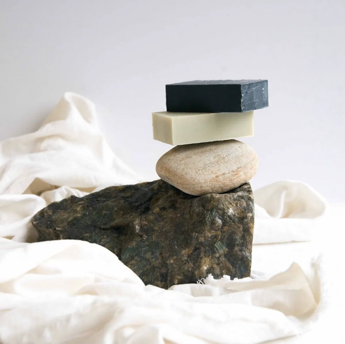 Stacked sculptural composition featuring rough stone, ceramic, wooden blocks, and metal pieces balanced on white fabric by Rebecca Johnson.