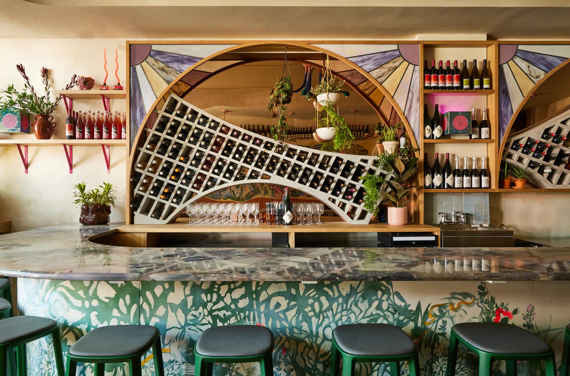 Modern bar interior with curved wooden archway, green patterned wallpaper, black seating, and tiered shelving displaying bottles and plants by Rebecca Johnson.