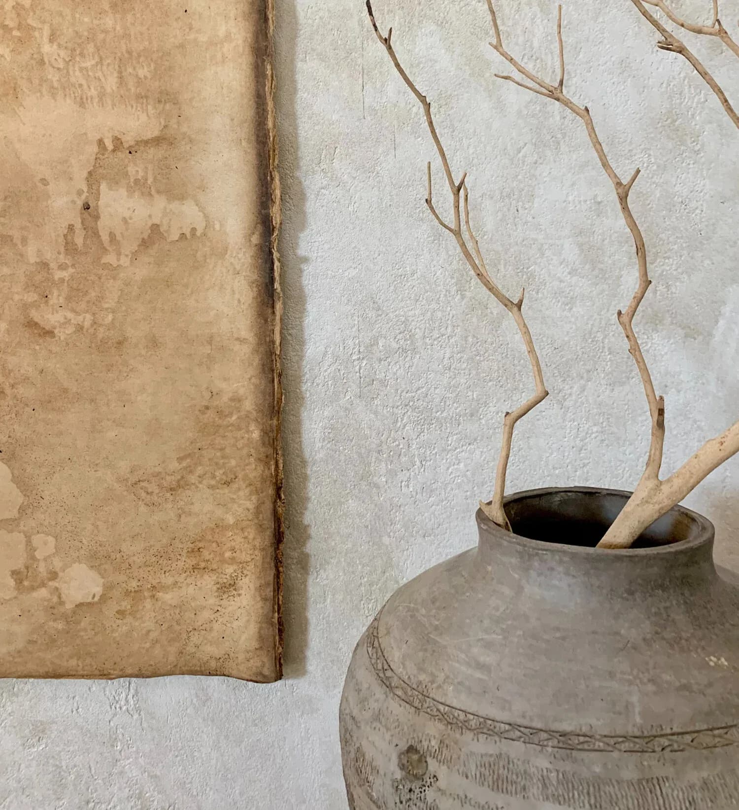 Minimalist interior detail featuring dried branches in a weathered ceramic vessel against textured plaster walls in neutral tones, by Rebecka Hekmat.