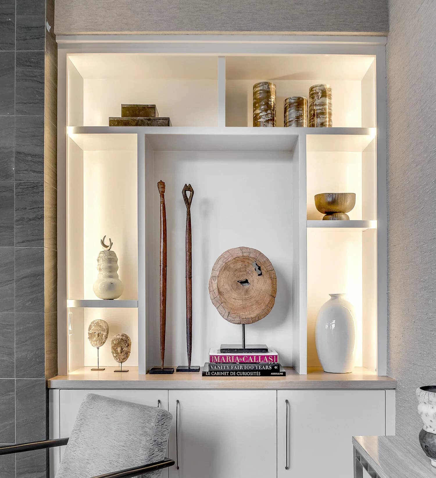 Built-in shelving display with warm lighting, featuring curated objects including wooden vessels, brass bowls, and decorative sculptures on white shelves, by Rebecka Hekmat.