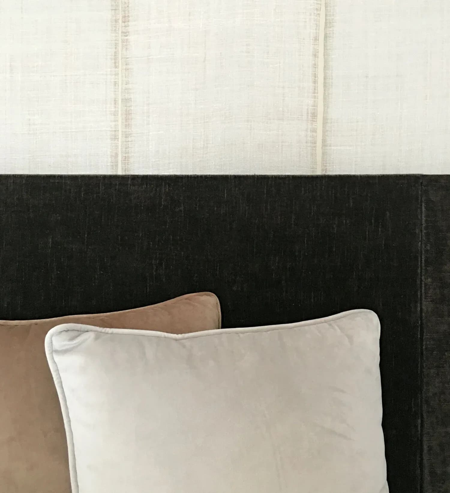 Minimalist bedroom detail showing a white pillow and taupe cushion against a black upholstered headboard with light cream wallpaper above, by Rebecka Hekmat.