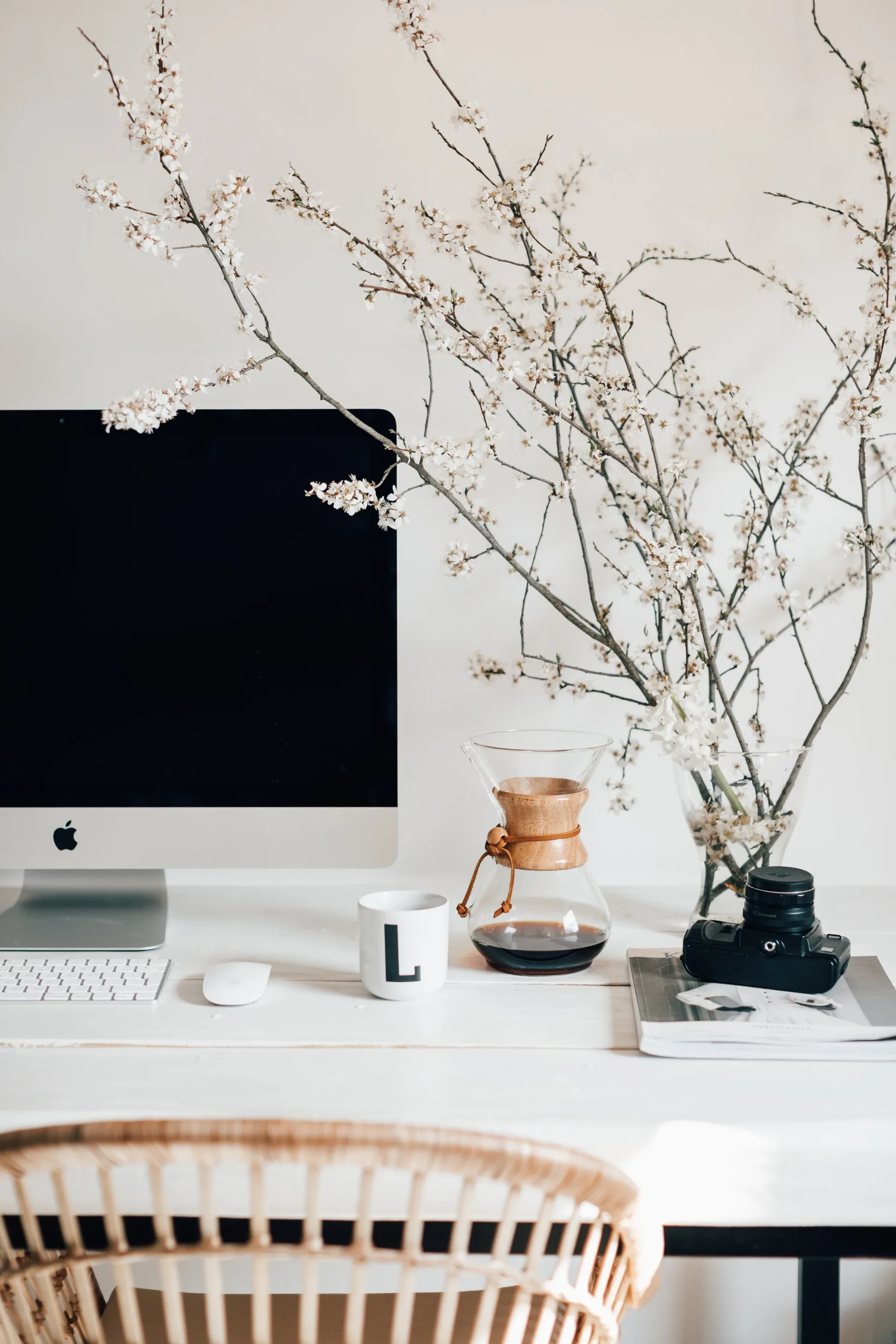 Minimalist workspace with white desk, potted dried branches arrangement, and Apple monitor, designed by Rebecka Hekmat.