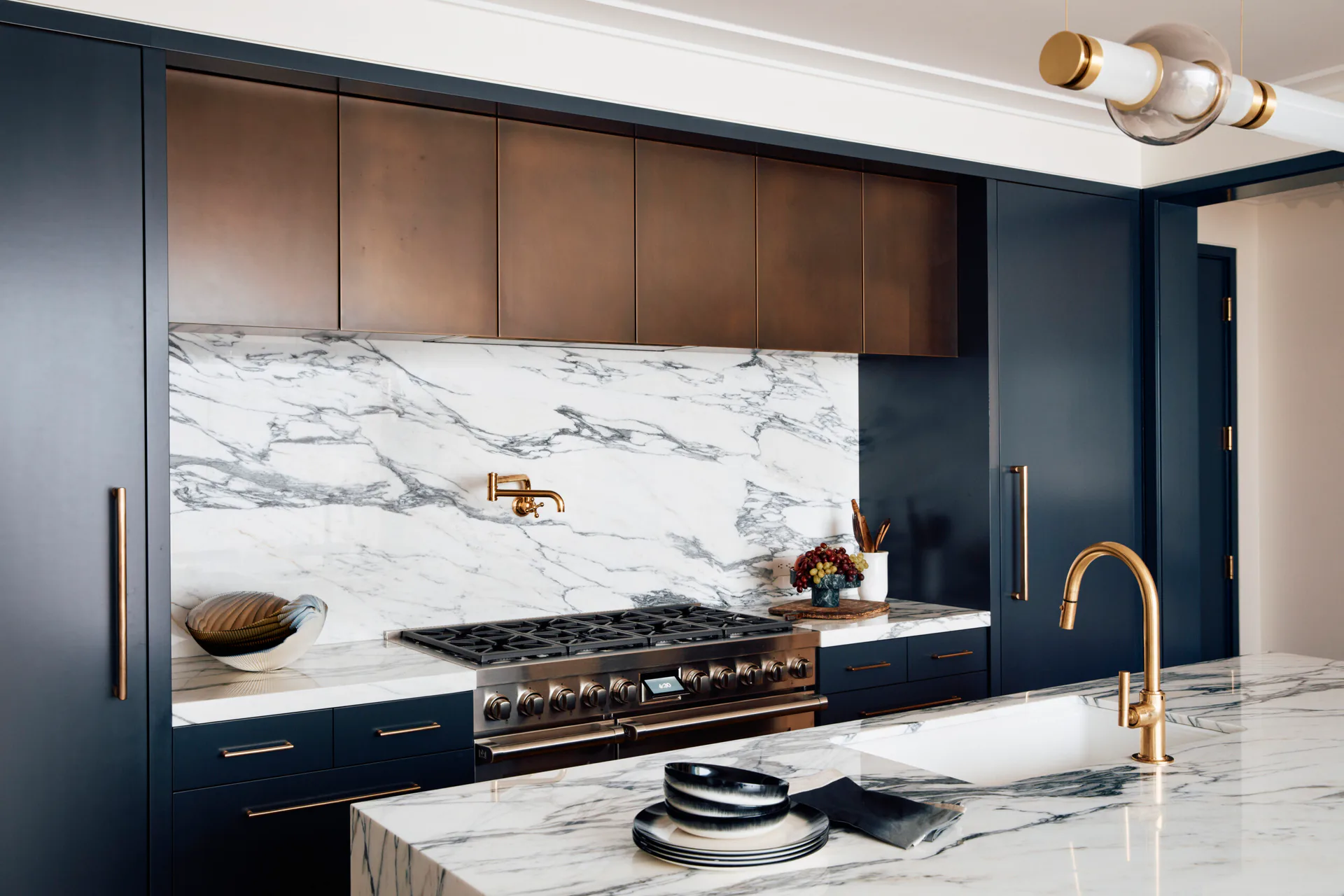 Modern kitchen with dark navy cabinetry, marble backsplash and countertops, brass fixtures, and a chef's range by Regina Garcia-marin.