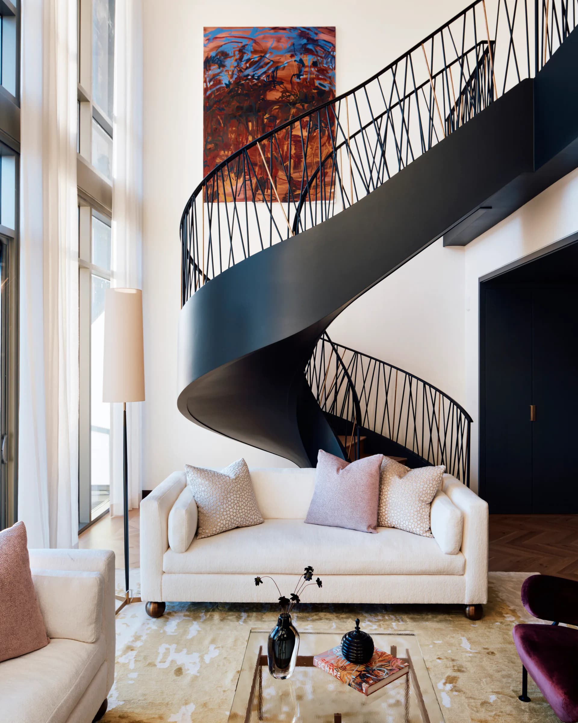 Modern loft interior featuring a dramatic black spiral staircase, cream sectional sofa with pink pillows, abstract red artwork, and high ceilings by Regina Garcia-marin.