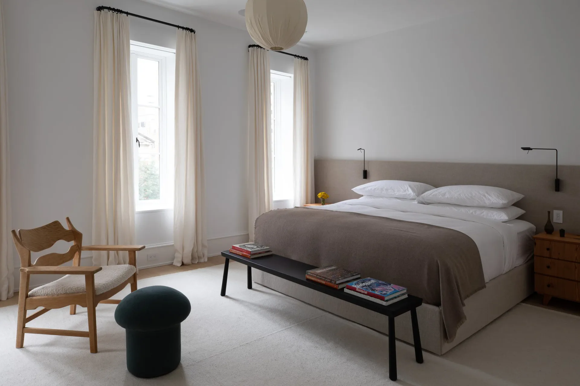 Minimalist bedroom with gray upholstered bed, wooden nightstand, natural light from tall windows with cream curtains, wooden chair, and black bench by Reinaldo Leandro.