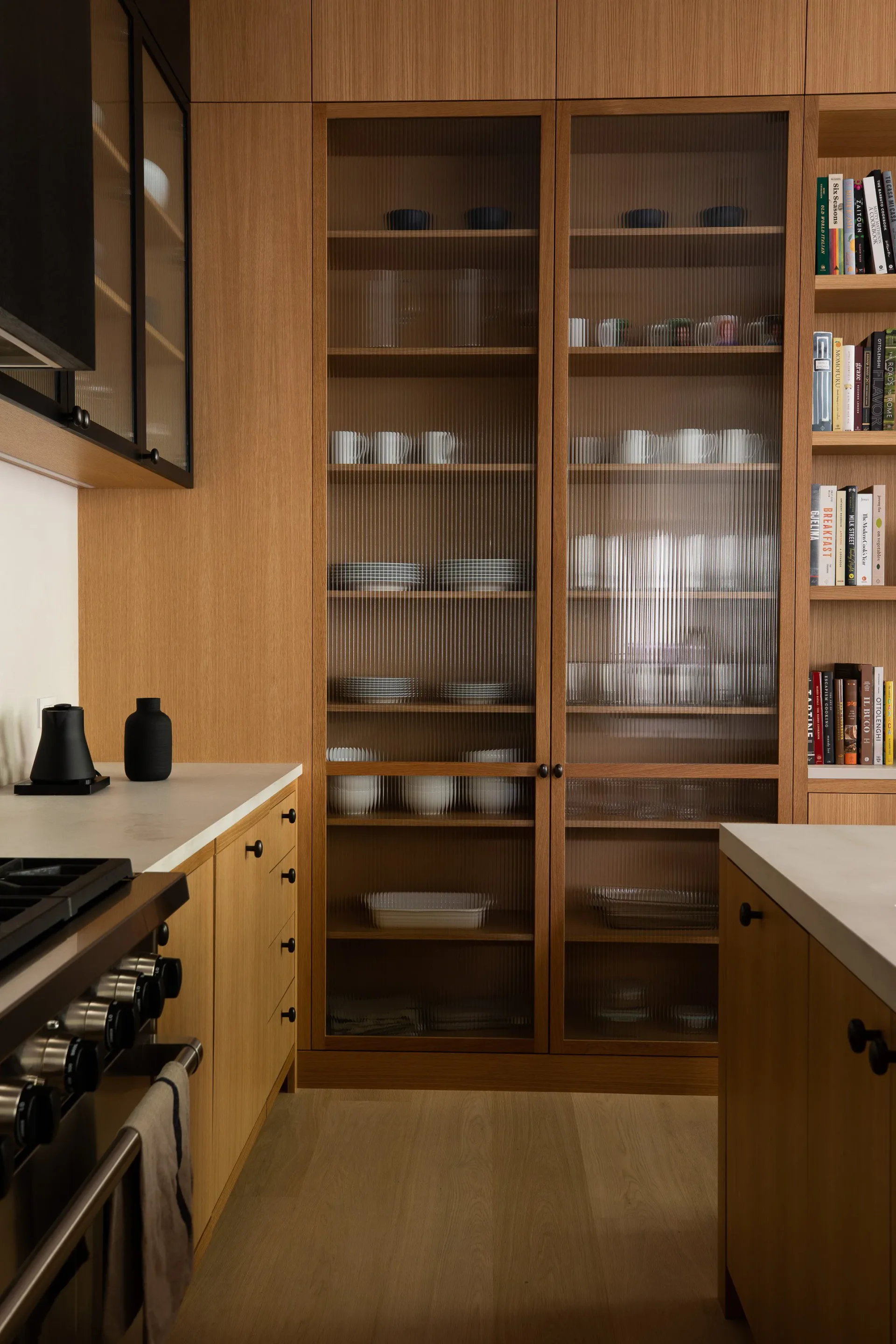 Modern kitchen with warm wood cabinetry, glass-fronted upper cabinets displaying white dishware, white quartz countertops, and integrated appliances, designed by Reinaldo Leandro.