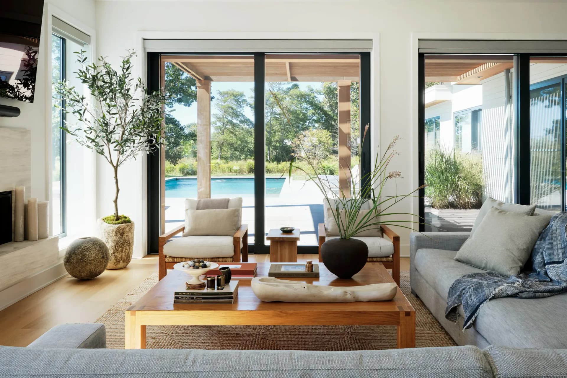 Bright contemporary living room with wooden coffee table, gray seating, large glazed doors opening to pool view, natural materials and plants throughout, by Remi Berger.
