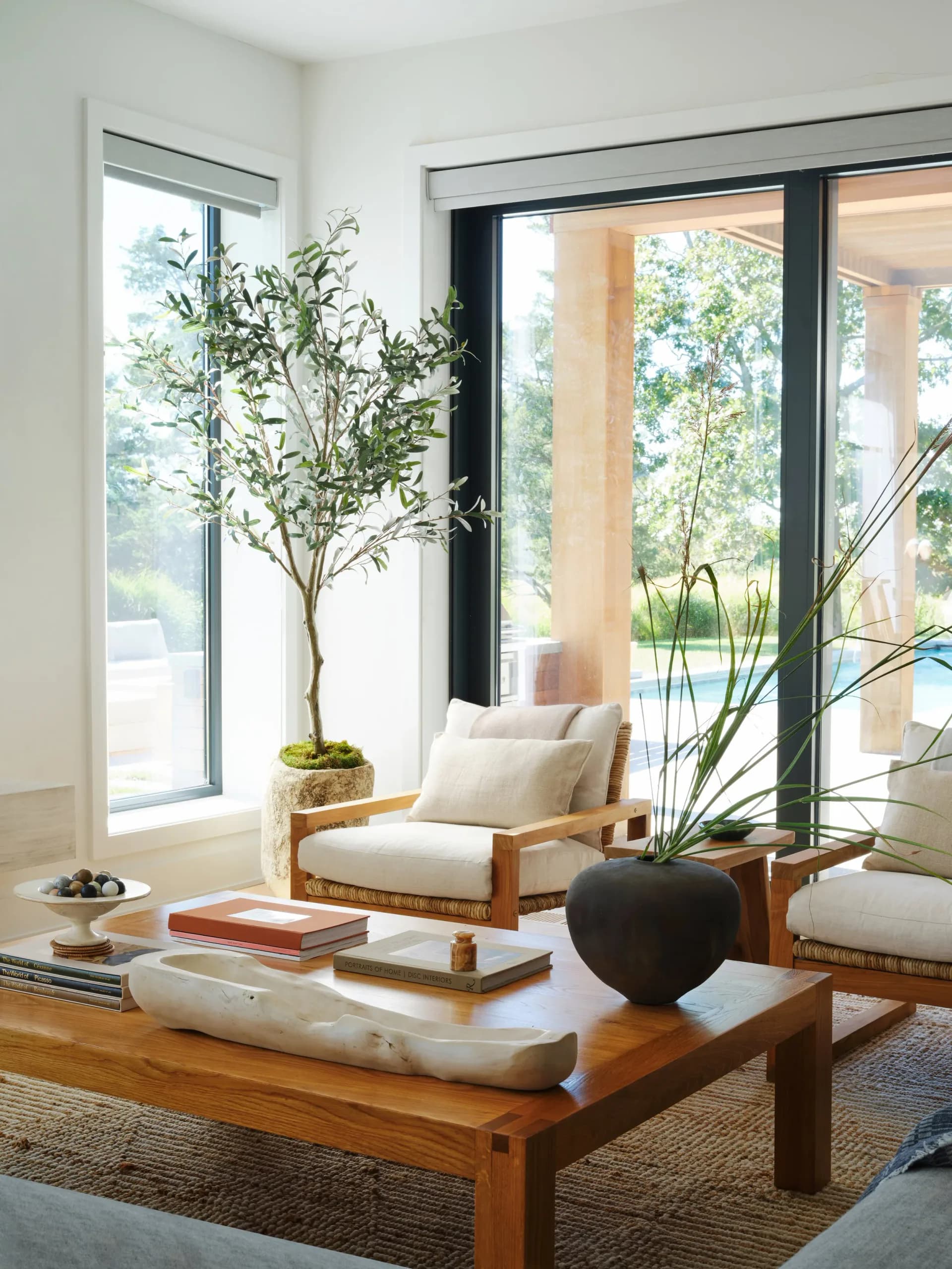 Minimalist living room with wooden furniture, cream cushions, potted plants, and expansive windows overlooking nature by Remi Berger.