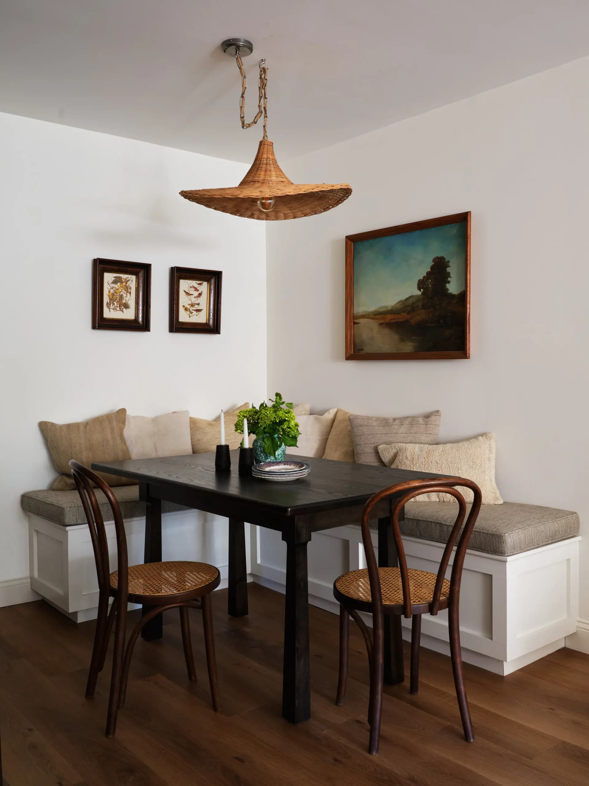 Dining nook with built-in banquette seating, black table, bentwood chairs, woven pendant light, and landscape painting by Remi Berger.