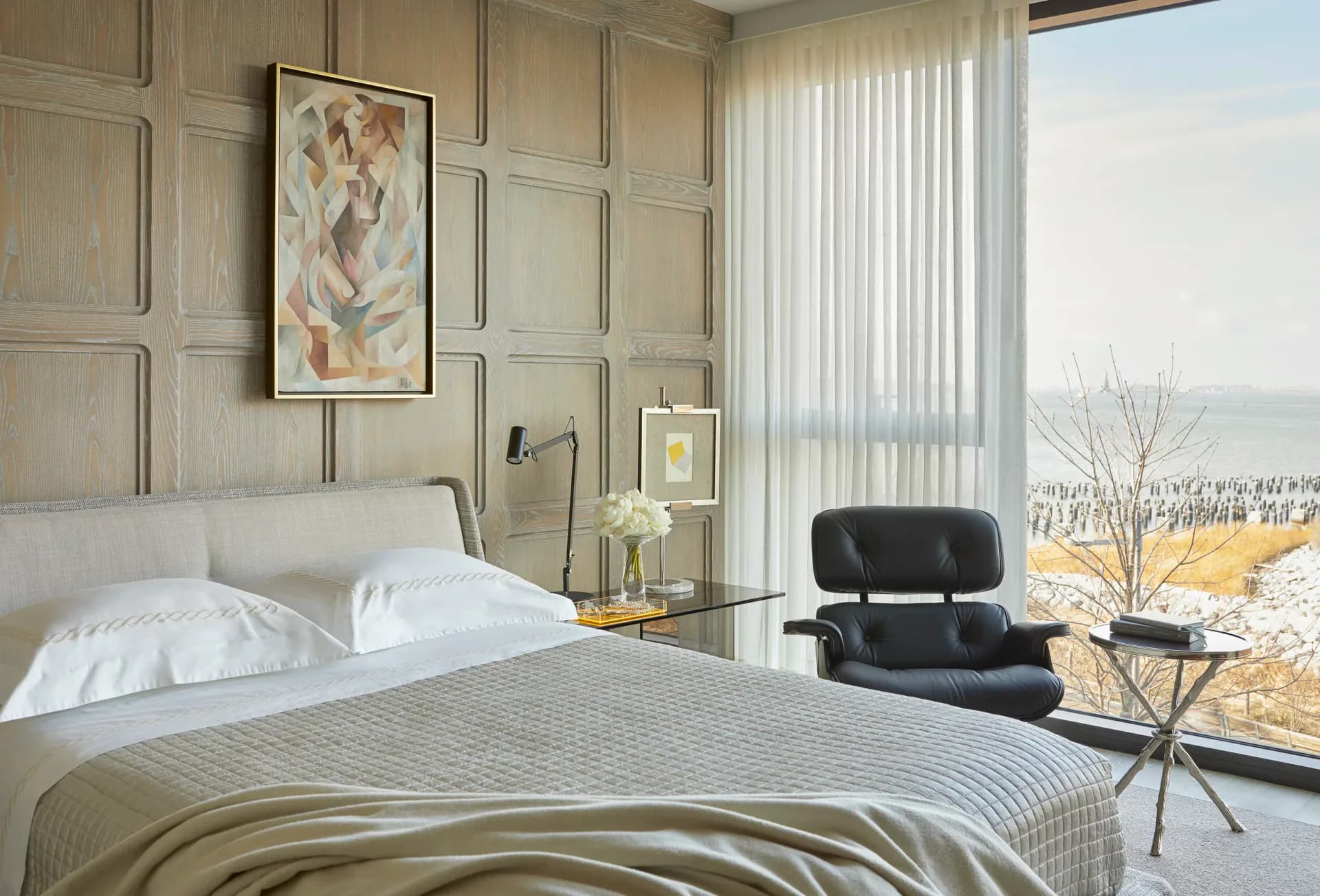 Serene coastal bedroom with neutral palette, paneled walls, white bedding, black lounge chair, and expansive ocean views by Remigio Gudin.