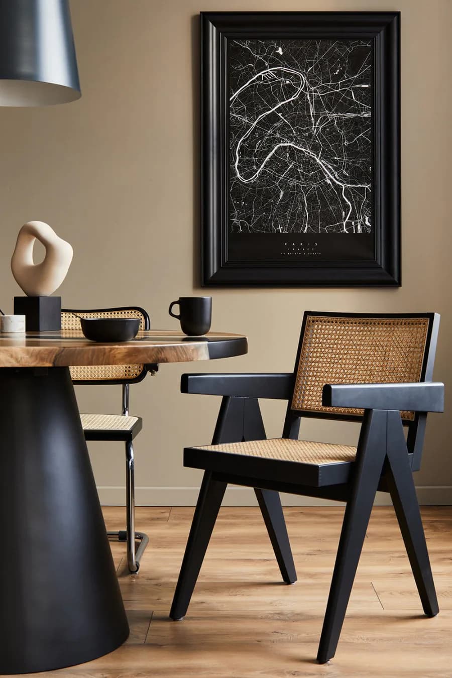 Modern minimalist interior featuring a black pedestal table with wooden top, caned chair with black frame, beige walls, and abstract map artwork by Remy Maurice.