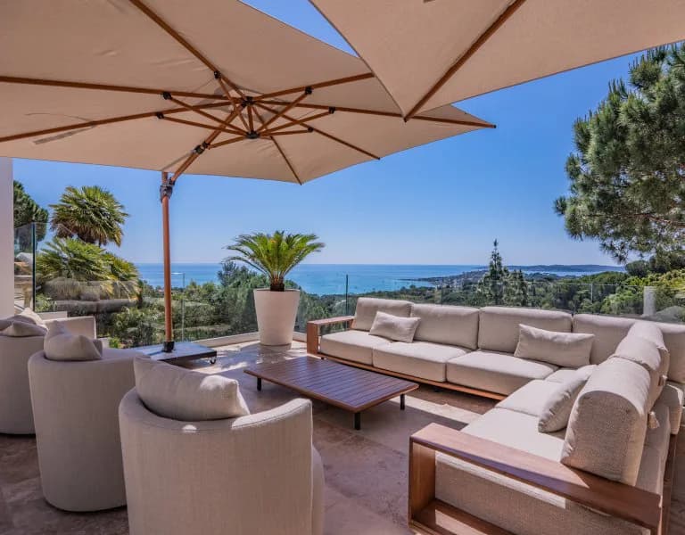 Luxury Mediterranean terrace with neutral upholstered seating, wooden umbrellas, sea views, and palm trees, designed by Remy Maurice.