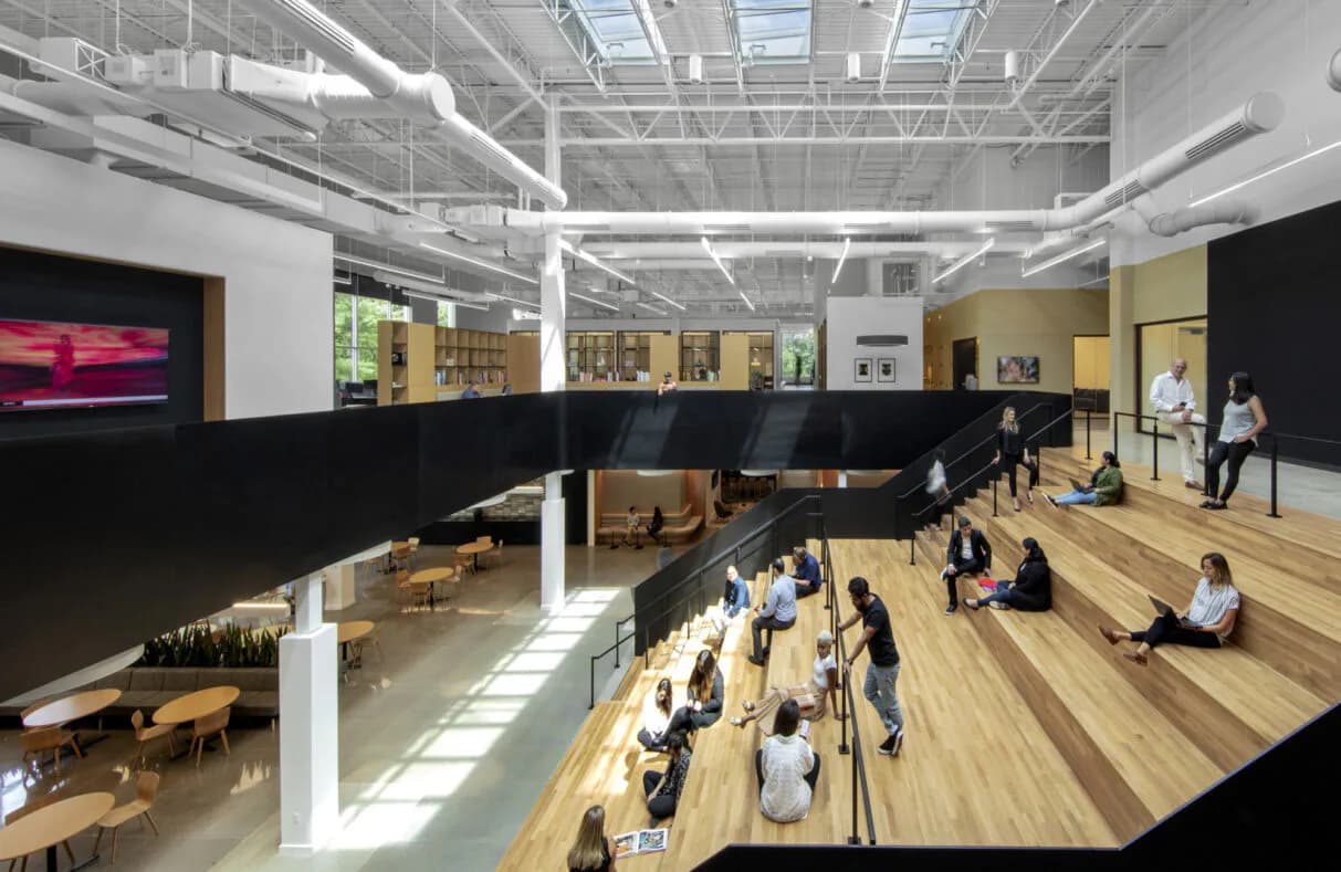 Modern open-plan interior with wooden flooring, white structural columns, and yellow-toned upper level featuring people in motion, designed by Rhea Thomas.