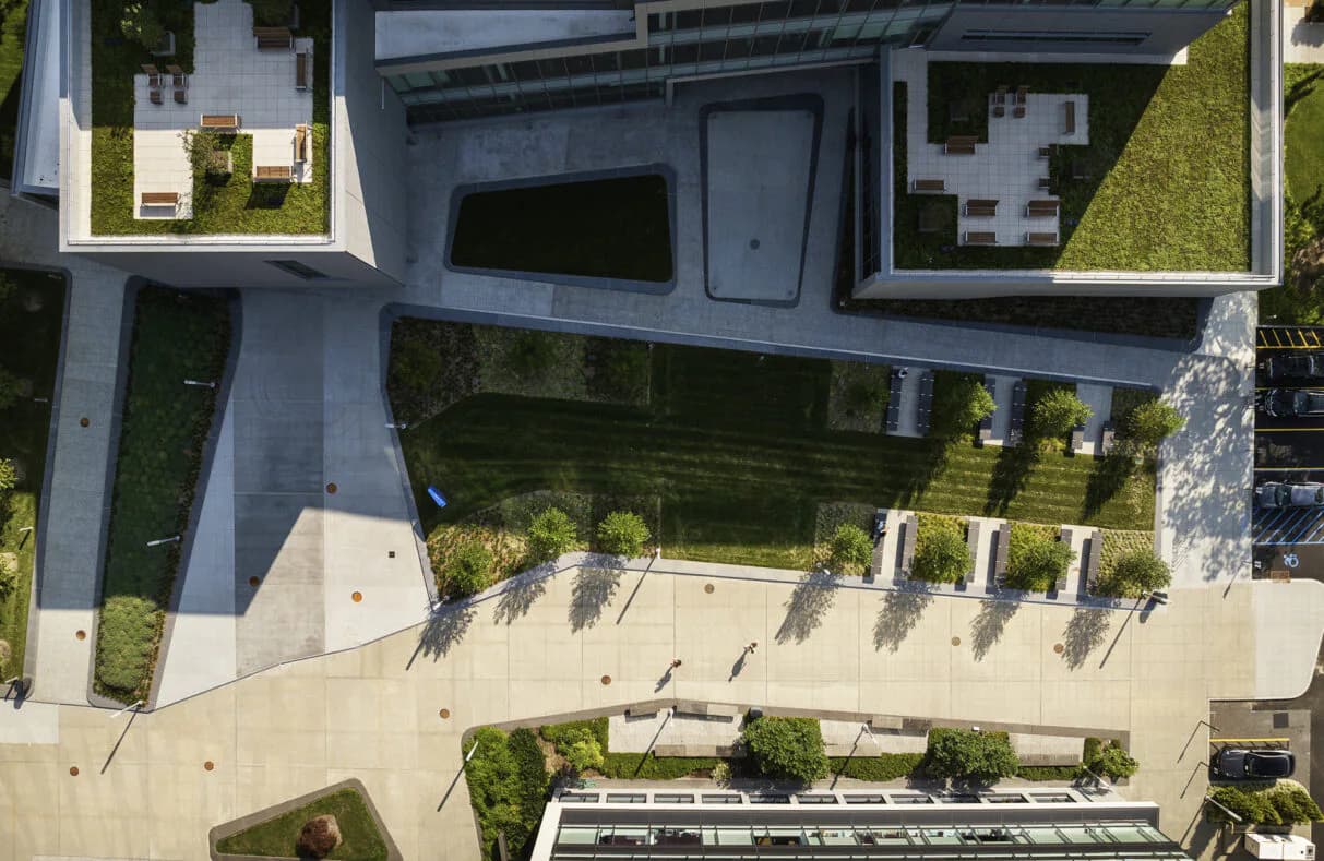 Aerial view of a modern courtyard with concrete paving, integrated planting beds, and contemporary architecture surrounding a green space by Rhea Thomas.