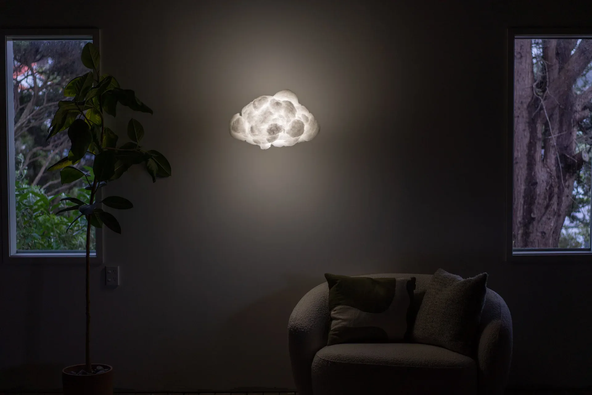 Dark minimalist interior with glowing cloud-shaped light fixture suspended above an olive green armchair, flanked by windows with nature views, by Richard Clarkson.