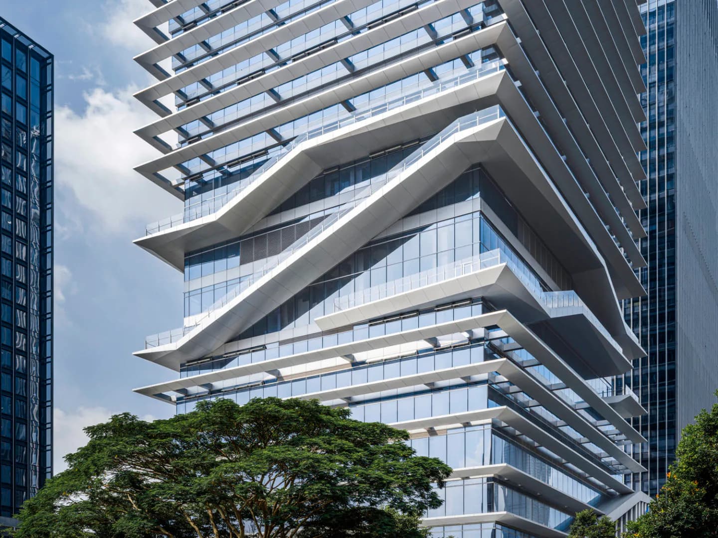 Modern curved residential tower with stacked white balconies and extensive glazing, rising above green trees against a blue sky by Richard Olcott.
