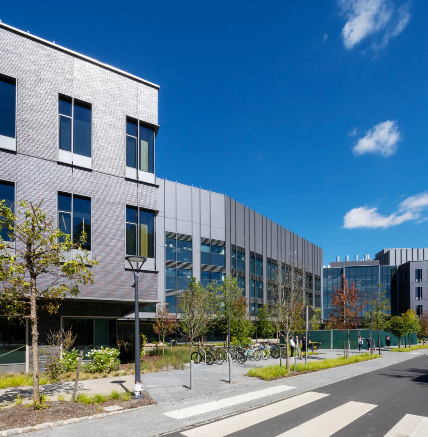 Contemporary mixed-use building with white brick facade, extensive glazing, and integrated landscape featuring young trees and bicycle parking by Richard Olcott.
