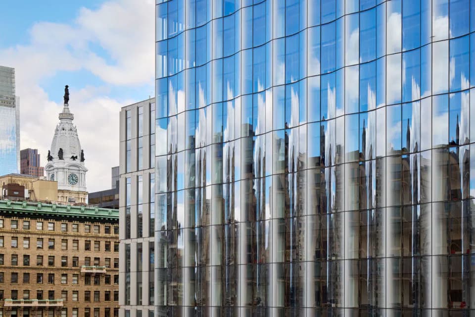 Modern glass-facade high-rise with vertical ribbed metallic panels reflects historic yellow stone buildings and a clock tower, designed by Richard Olcott.