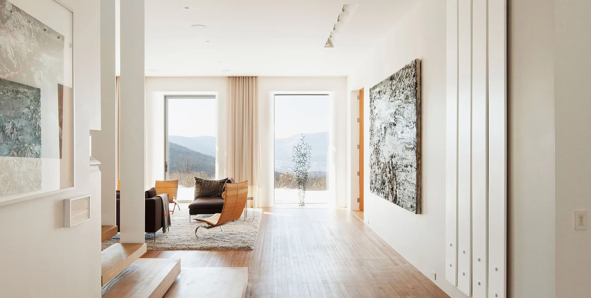 Modern minimalist interior with white walls, light wood flooring, orange accent chair, and mountain views through large windows, designed by Rick Cook.
