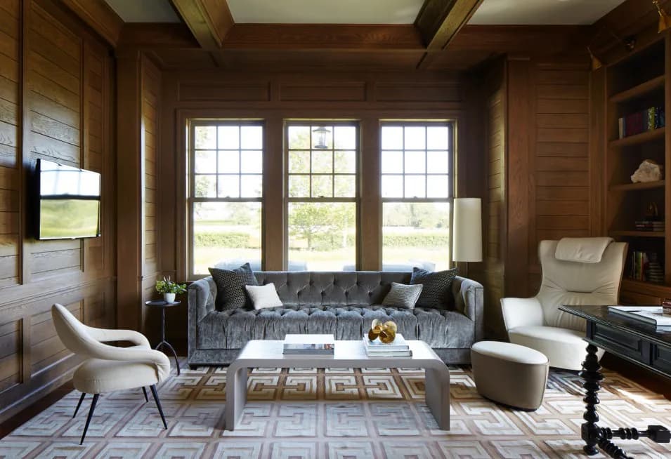 Luxurious living room with dark wood paneling, large multi-pane windows, gray tufted sofa, cream accent chairs, geometric patterned rug, and built-in shelving by Rinat Lavi.