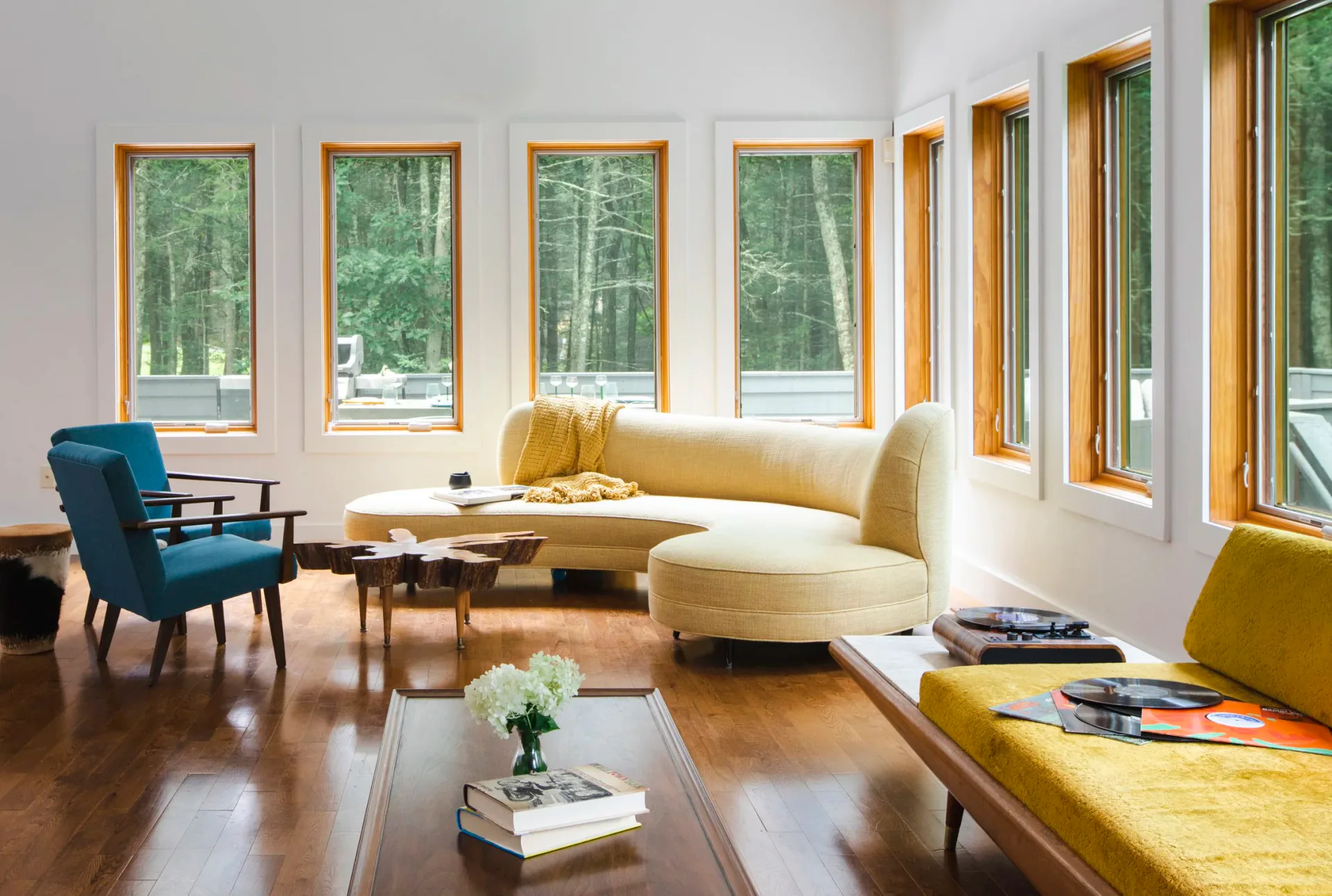 Bright modern living room with wood flooring, cream curved sofa, blue accent chair, and olive-yellow ottoman, lined with multiple large windows overlooking forest by Ritika Bhasin.