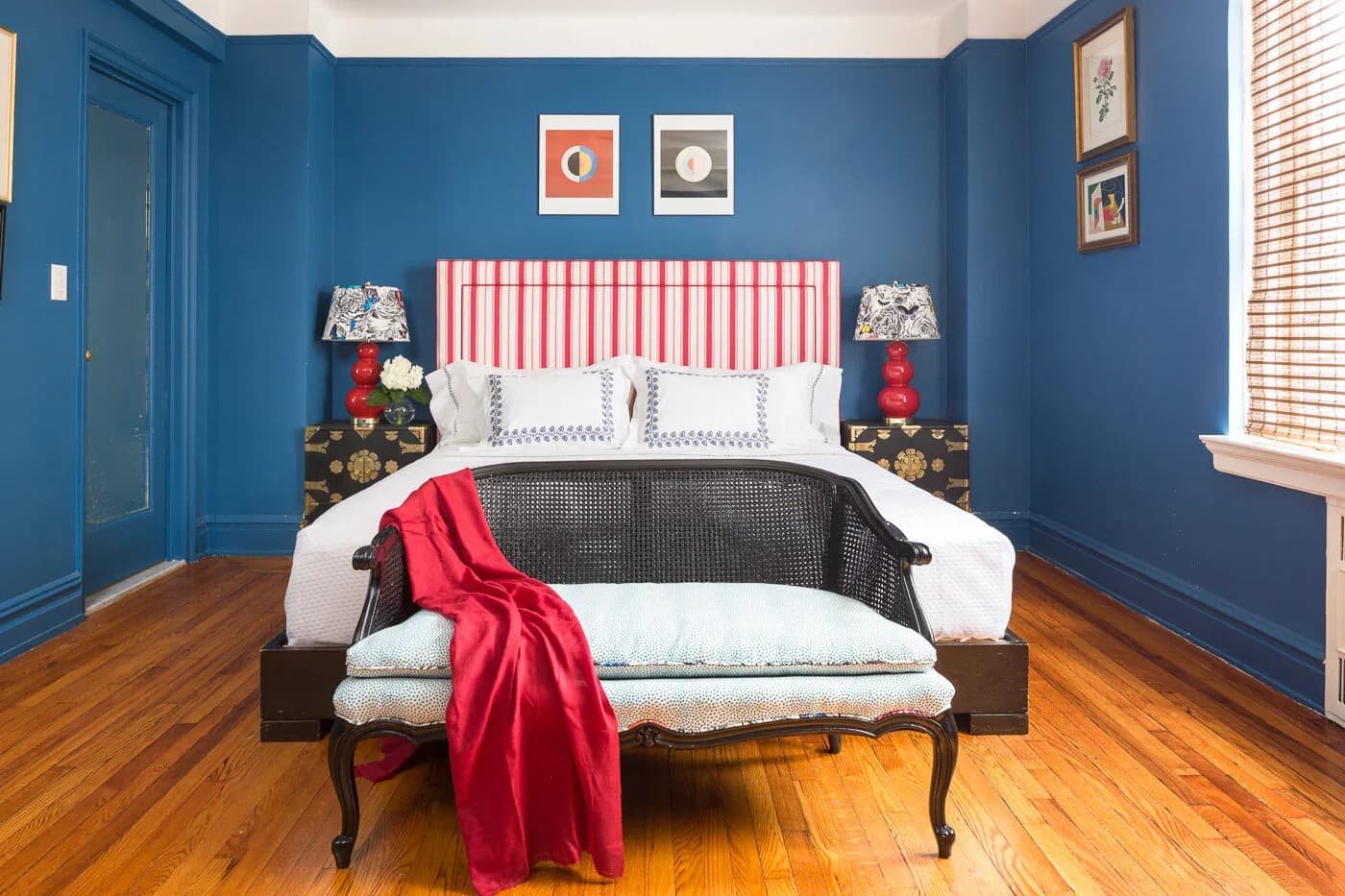 A bold blue bedroom with a red and white striped headboard, dark nightstands with lamps, hardwood floors, and decorative artwork above the bed by Ritika Bhasin.