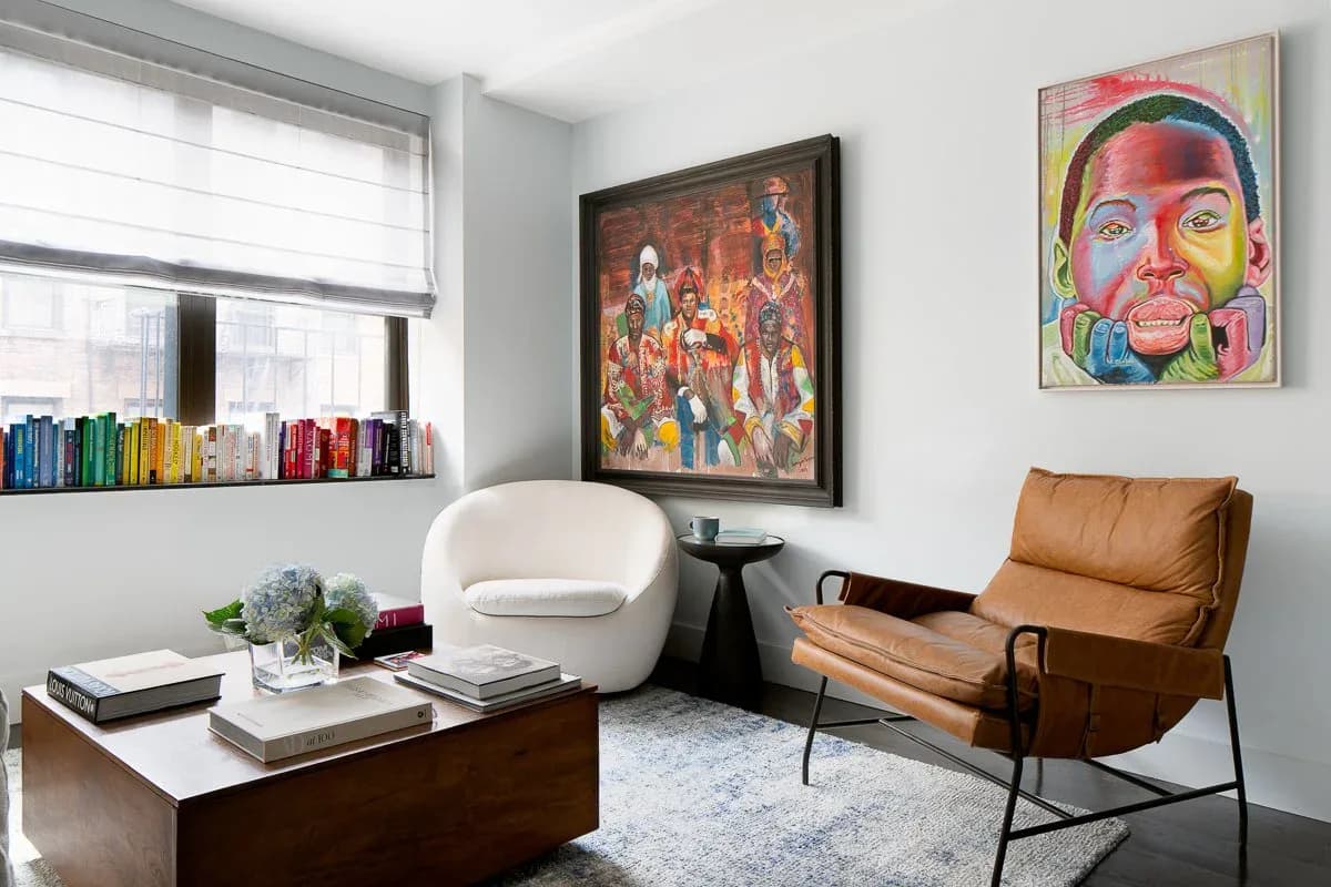 Contemporary living room with cream curved chair, cognac leather lounge seat, dark wood coffee table, colorful artwork, and natural light from large windows by Ritika Bhasin.