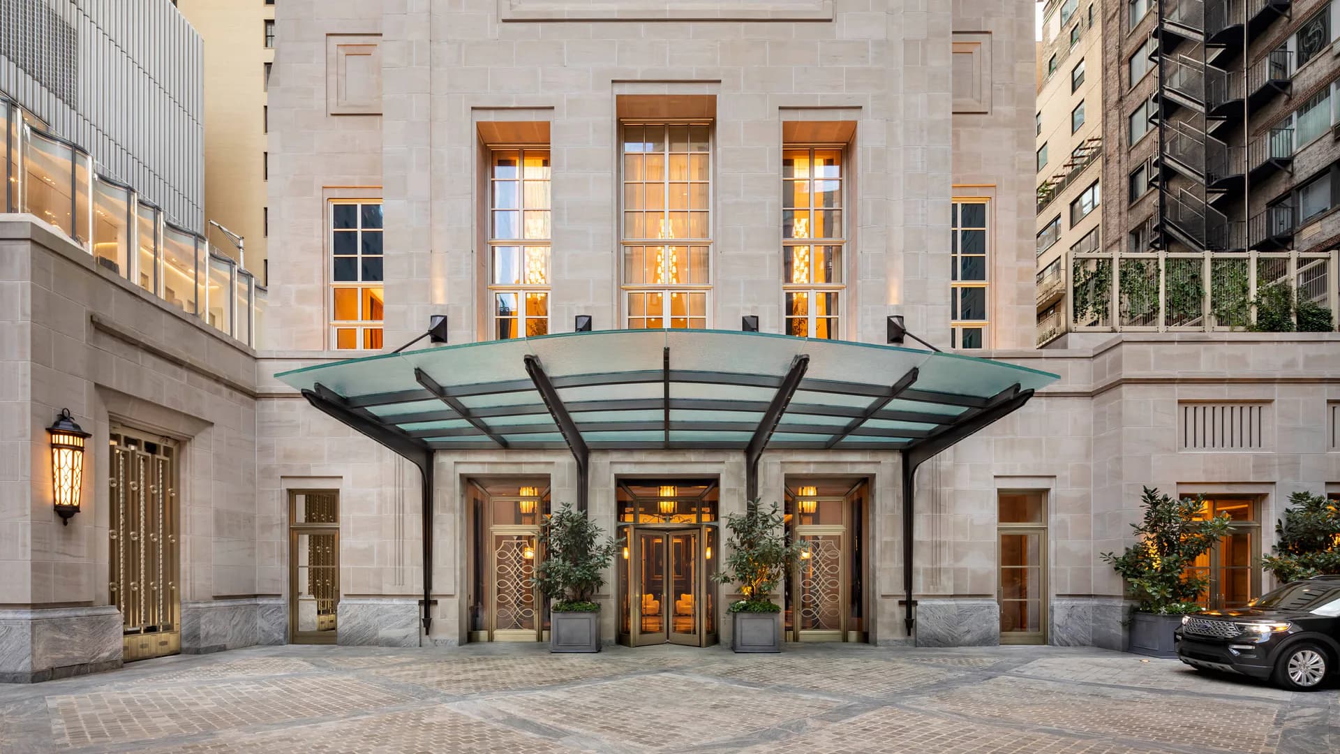 Modern urban courtyard entrance featuring a glass and steel canopy over illuminated glass doors, limestone facade, and manicured landscaping by Robert A.M. Stern.
