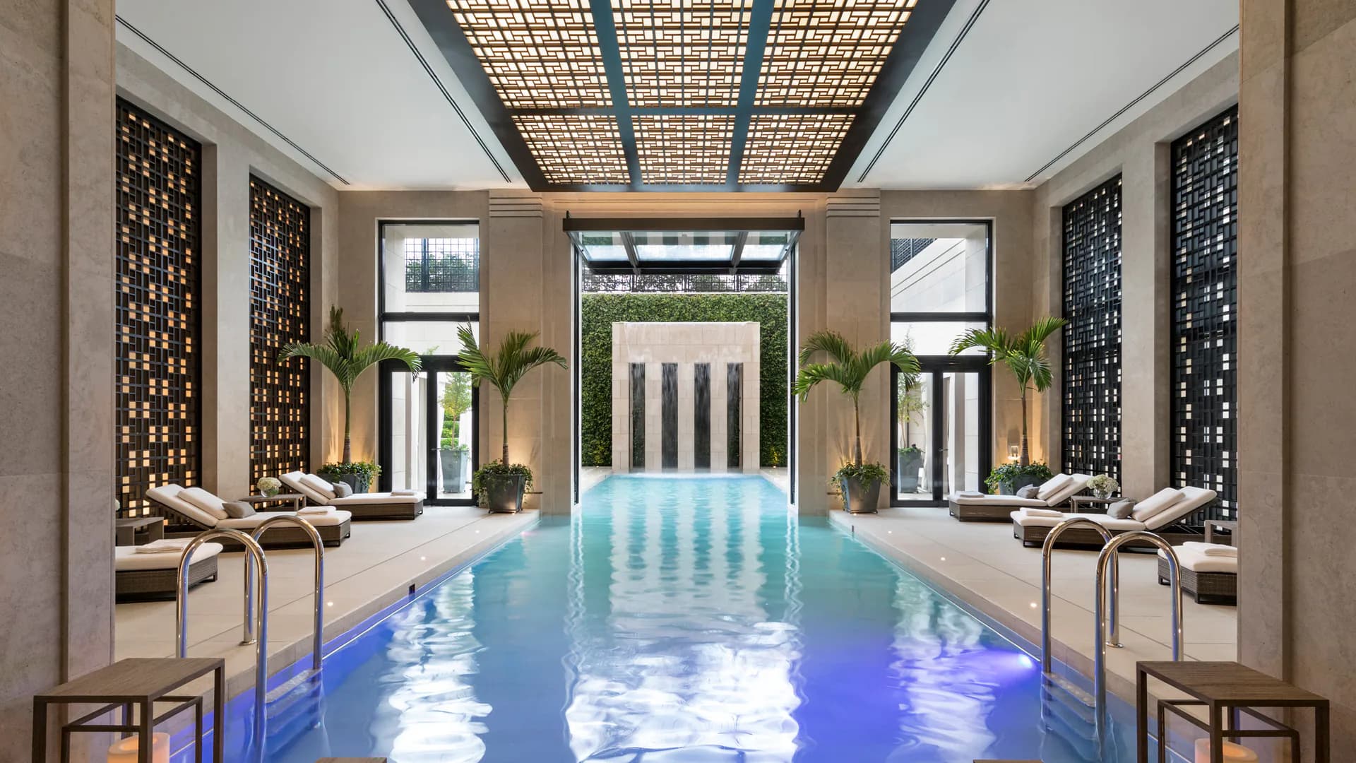 Luxurious indoor swimming pool with turquoise water, coffered ceiling with skylight, symmetrical loggias with columns and armchairs, potted palms, and geometric screen walls by Robert A.M. Stern.
