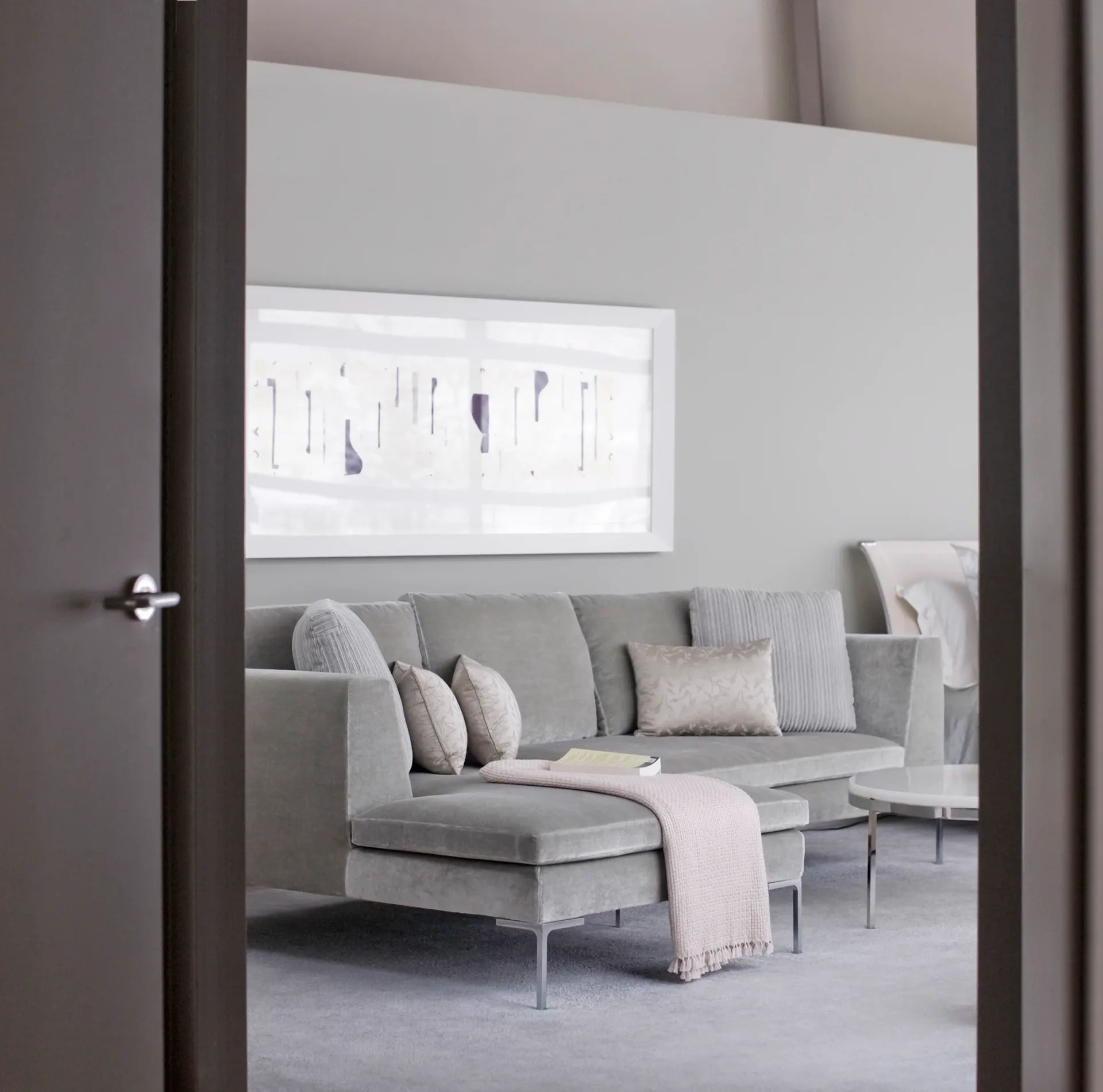 Modern living room with gray sectional sofa, layered pillows, and window with abstract artwork by Robert Nassar.