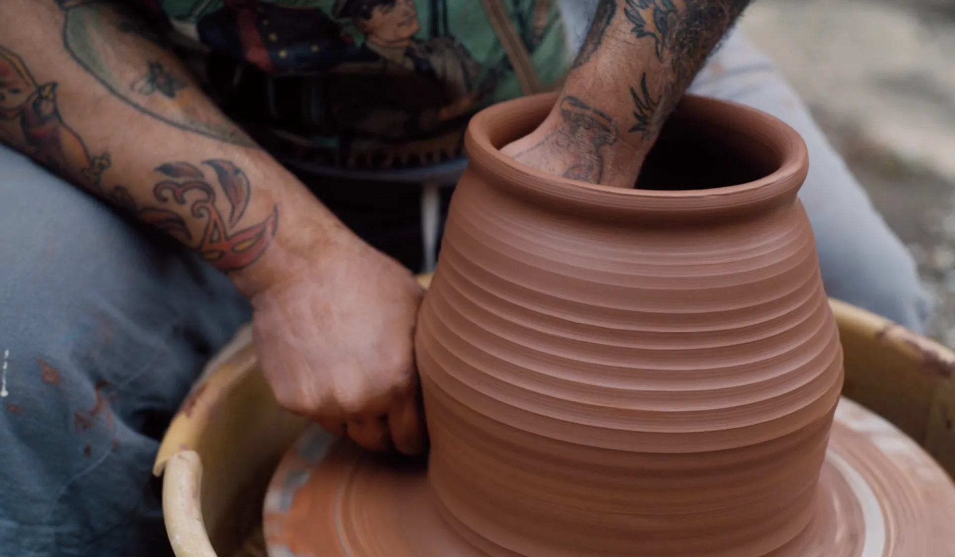 Hands shaping a terracotta ceramic vessel on a pottery wheel, showing textured horizontal ridges and natural clay tones by Roberto Lugo.