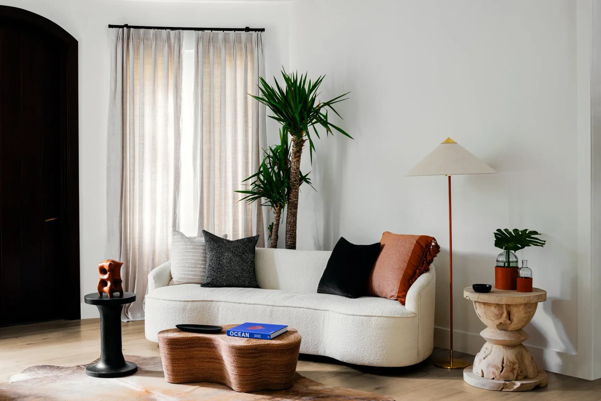 Minimalist living room with curved cream sofa, black and terracotta pillows, woven ottomans, potted plants, and floor lamp by Roberto Sulic.