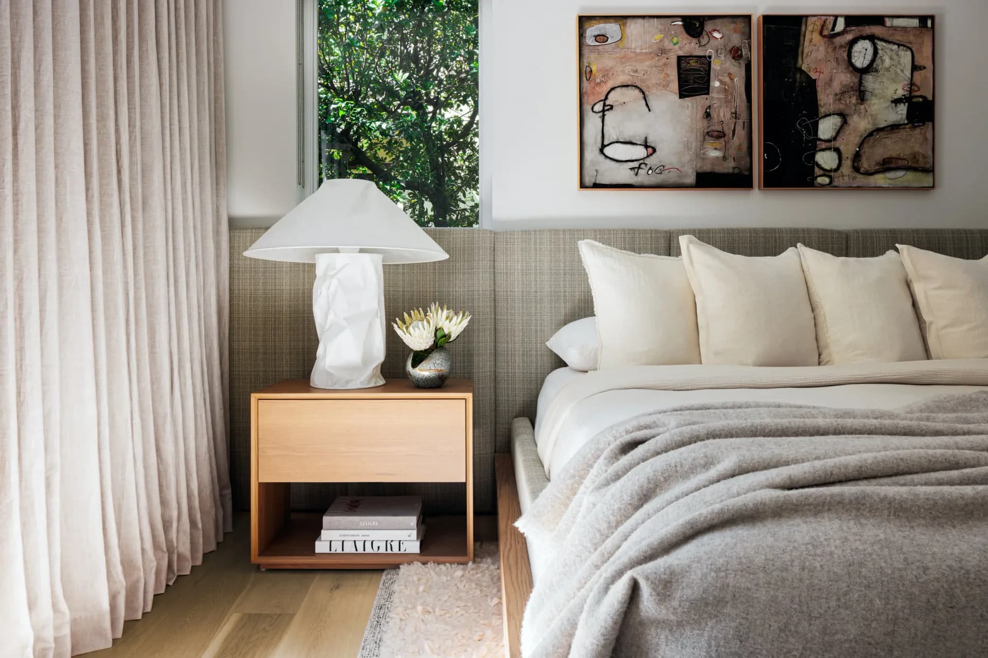 Serene modern bedroom with gray upholstered headboard, layered neutral bedding, wooden nightstand with white sculptural lamp, and floor-to-ceiling curtains by Roberto Sulic.