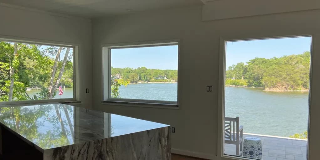Bright waterfront room with multiple large windows framing views of a lake and tree-lined shores, featuring modern interior design by Rosanna Trivilino.