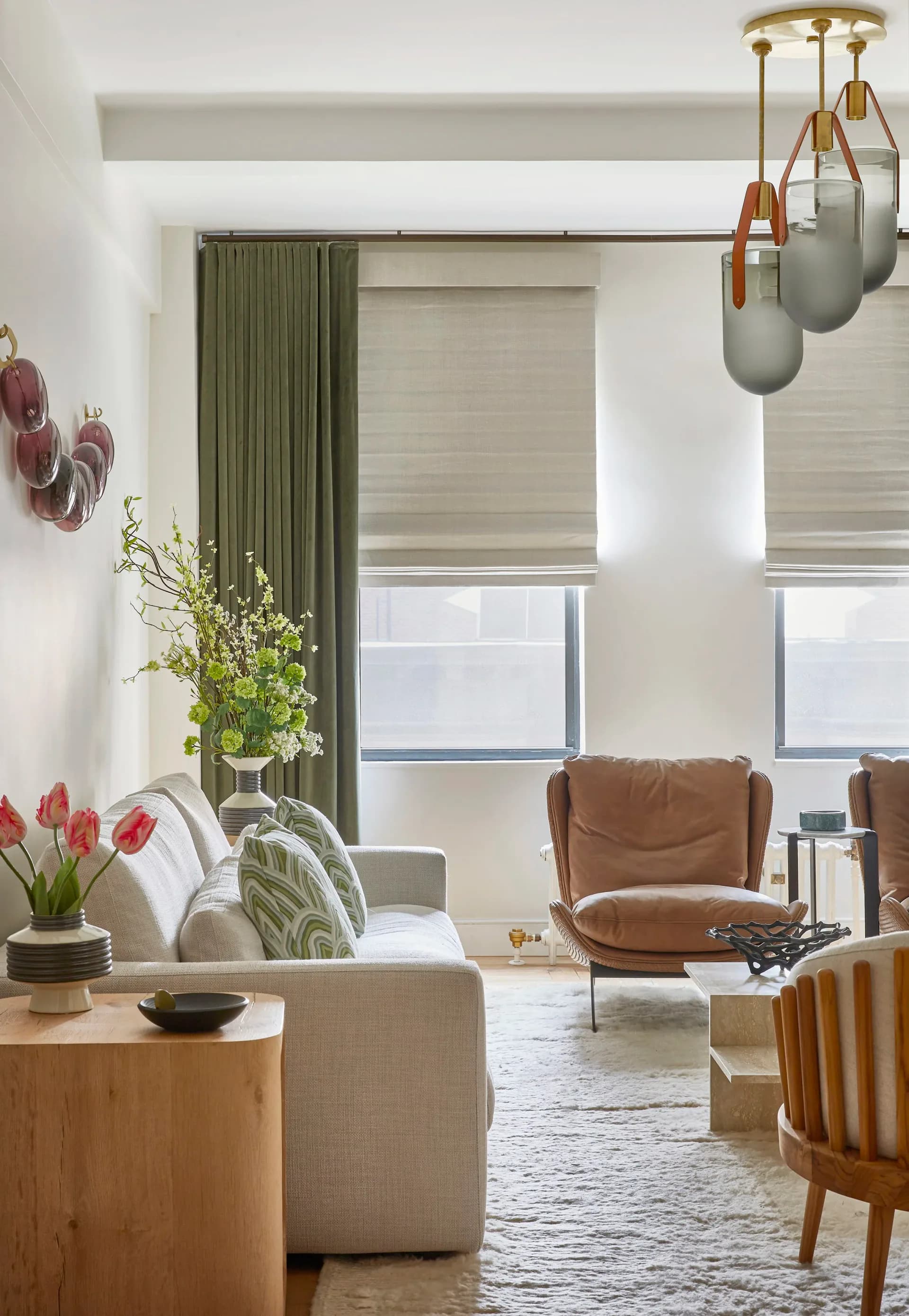 Modern living room with beige sectional, cognac leather chair, olive green curtains, pendant lighting, and natural wood accents by Ruchi Mohan.