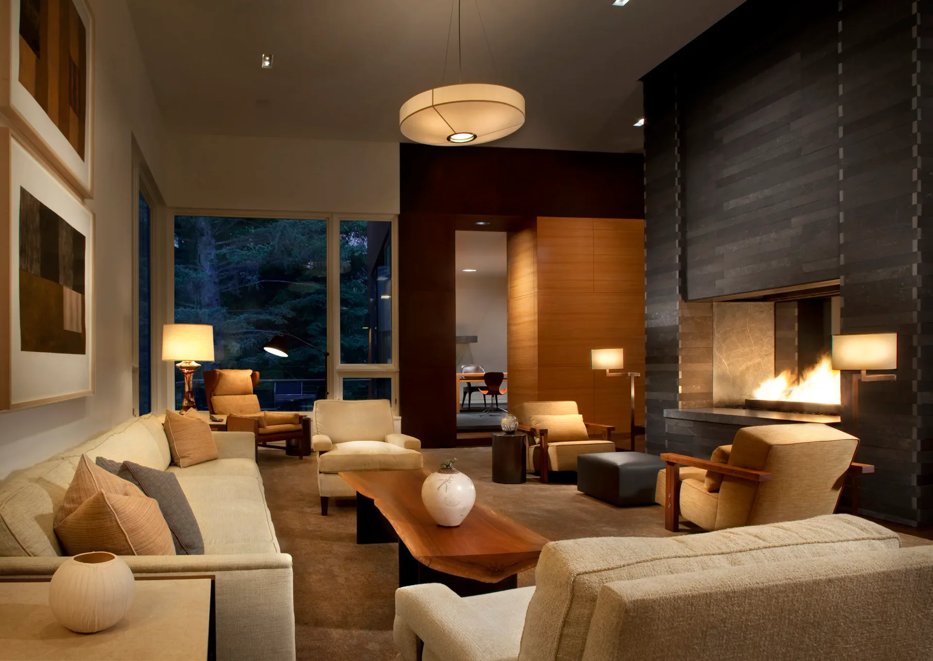 Contemporary living room with cream sofas, wooden coffee table, warm ambient lighting, and floor-to-ceiling windows overlooking evening landscape by Ruthie Osborne.