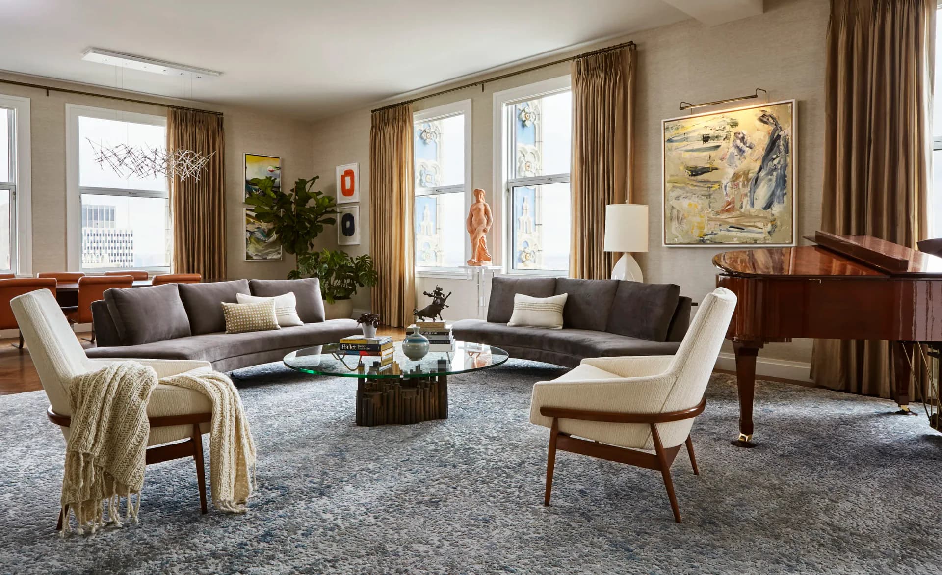Elegant living room with gray upholstered sectional, cream accent chairs, and wood furnishings arranged around a central table, featuring large windows with gold curtains and neutral palette by Ruthie Osborne.
