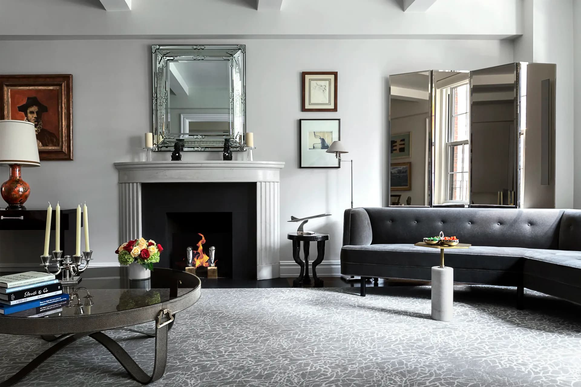 Modern living room with curved charcoal sofa, marble fireplace, mirrored mantel, geometric coffee table, and neutral palette by Ruthie Osborne.