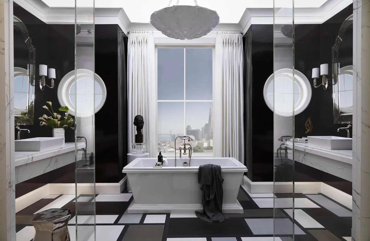 A luxury bathroom with symmetrical black marble walls, white marble vanities with circular mirrors, freestanding soaker tub, and floor-to-ceiling windows overlooking a city skyline by Ruthie Osborne.