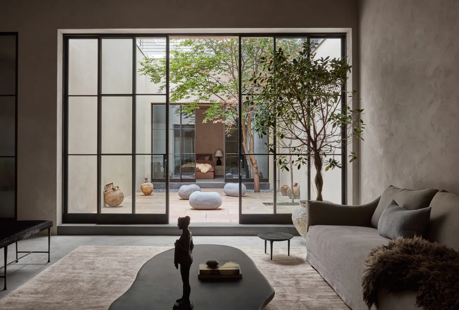 Modern living room with floor-to-ceiling gridded glass doors opening to a courtyard, featuring gray furnishings, polished concrete floors, and minimalist decor by Ryan Korban.