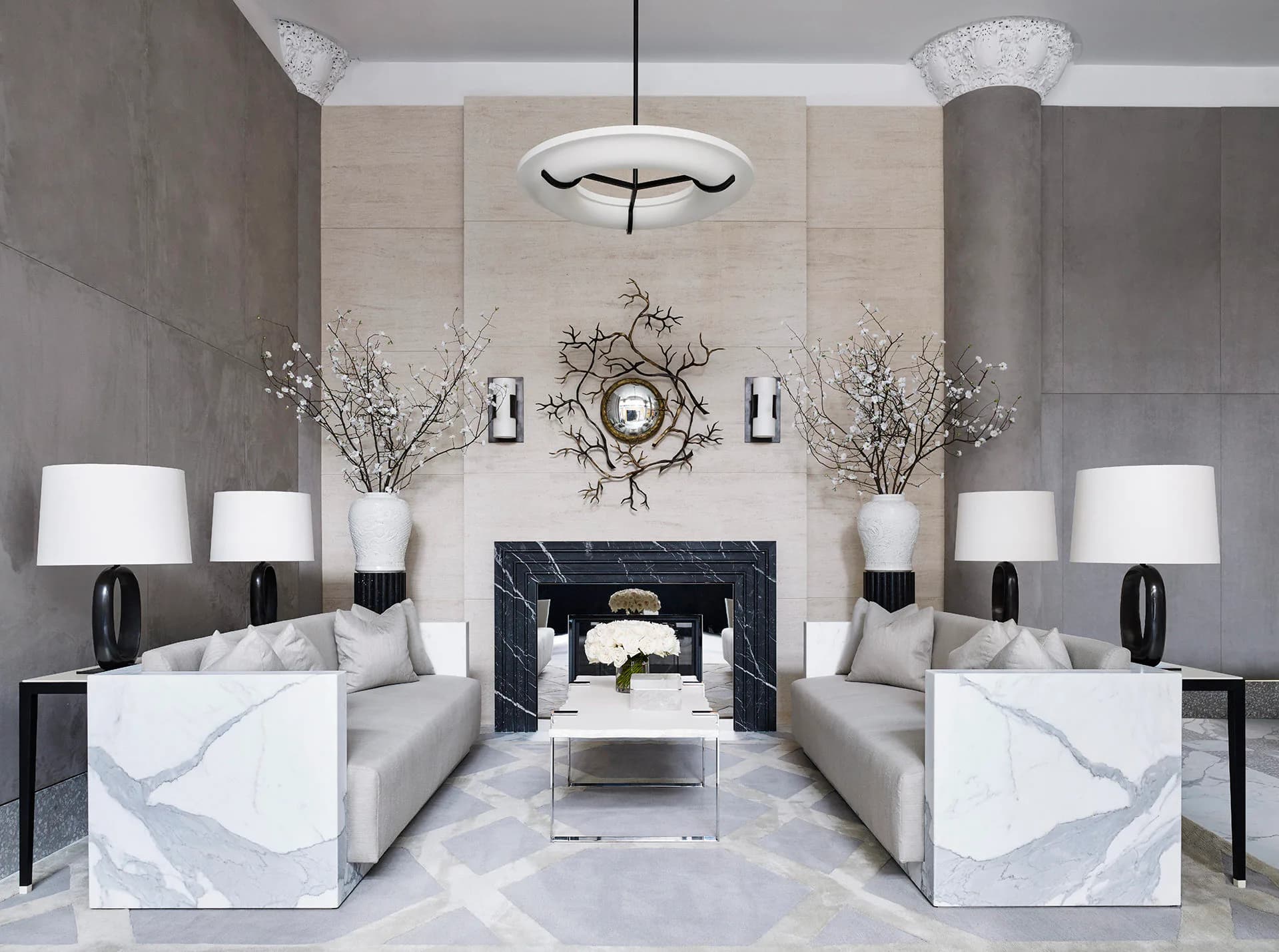 Symmetrical modern living room with dual white marble sofas, black fireplace, decorative branches, and pendant lighting by Ryan Korban.