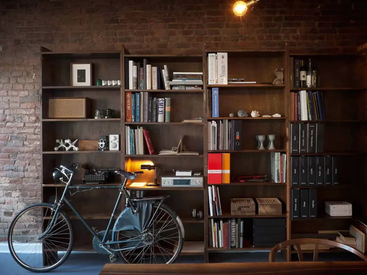 Dark wooden shelving unit filled with books, decor, and a blue bicycle, against exposed brick wall with warm lighting by Ryan Mahoney.