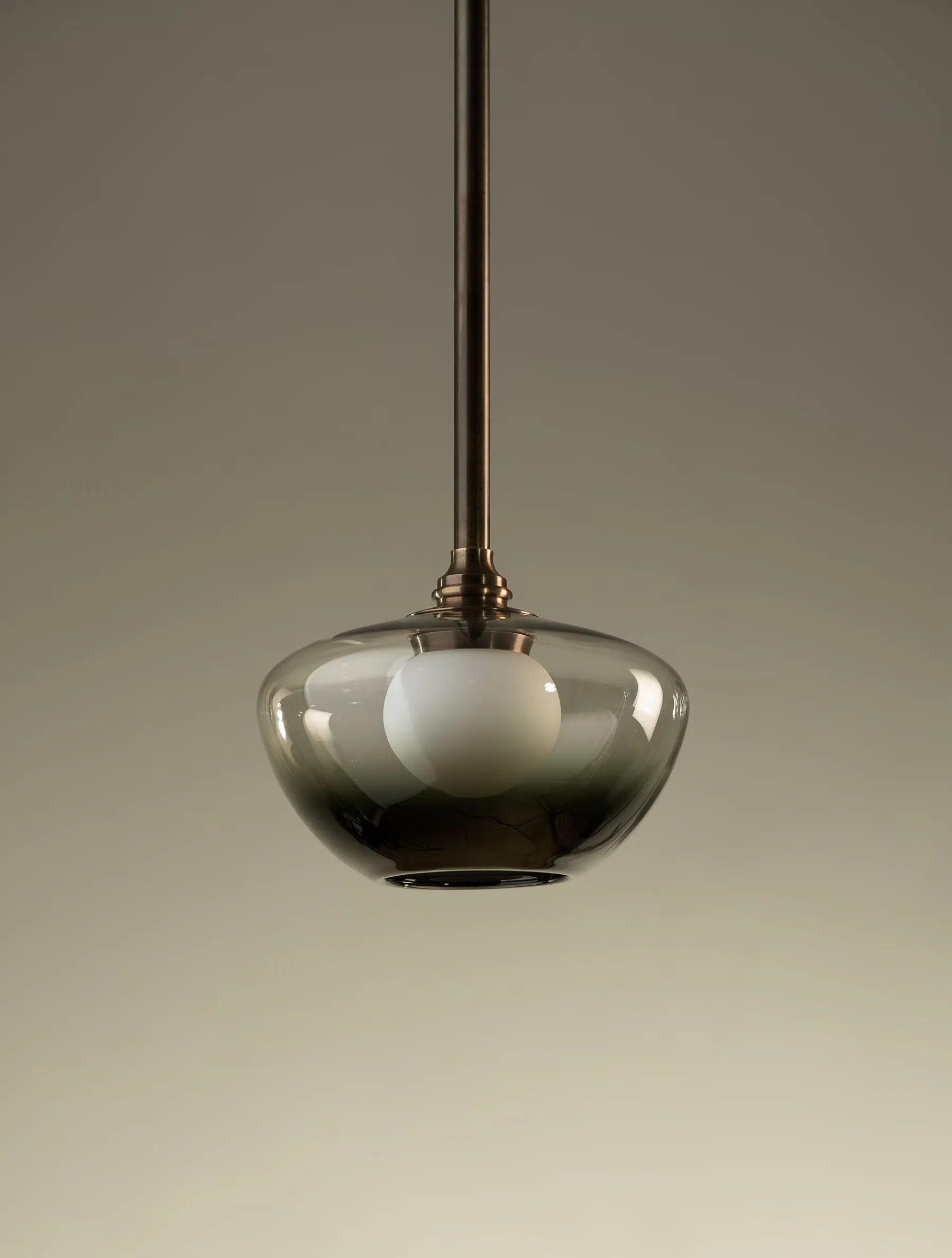 Modern pendant light with smoky grey glass dome and brass hardware suspended from rod, warm neutral background by Ryden Rizzo.