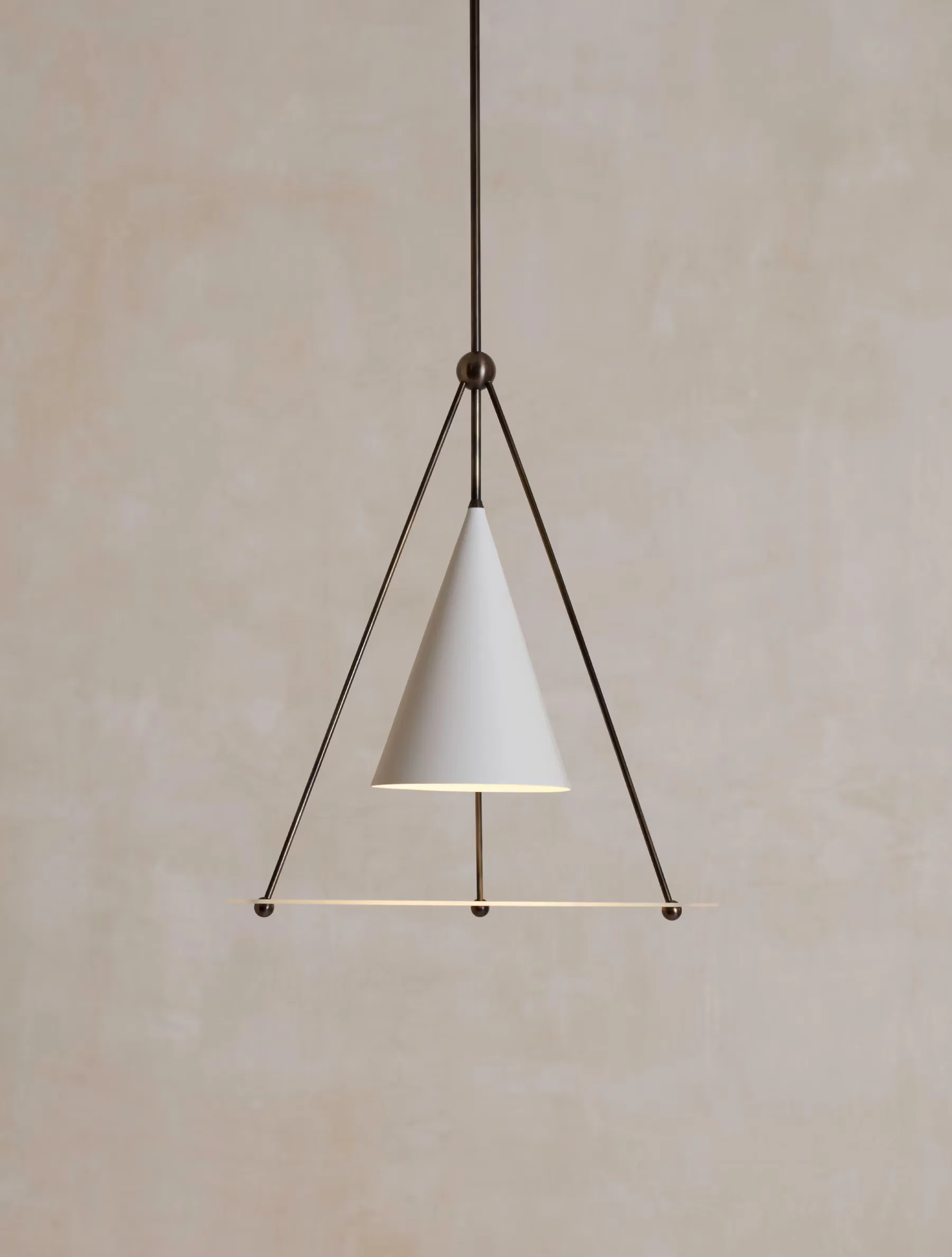 Minimalist pendant light with white cone shade suspended by metal tripod frame with bronze hardware and rod against neutral wall by Ryden Rizzo.